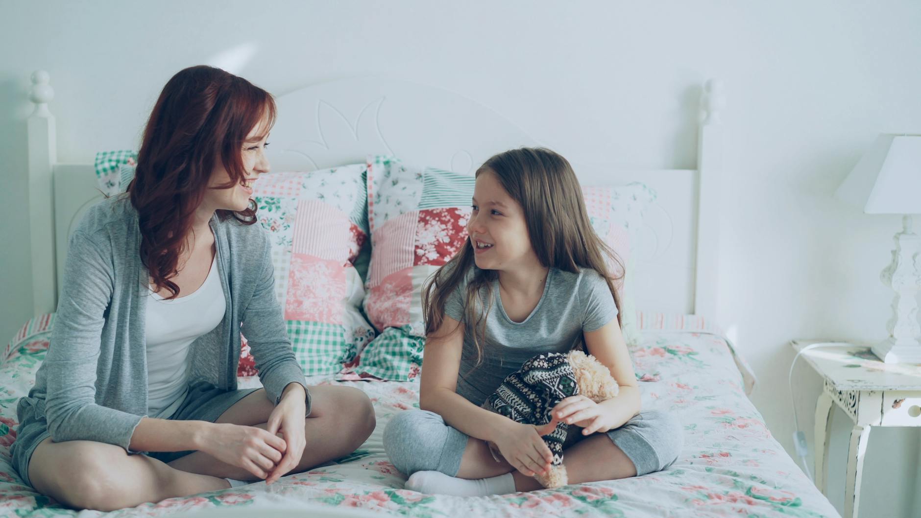 Uśmiechnięta mama z córką rozmawiają na łóżku w przytulnym pokoju